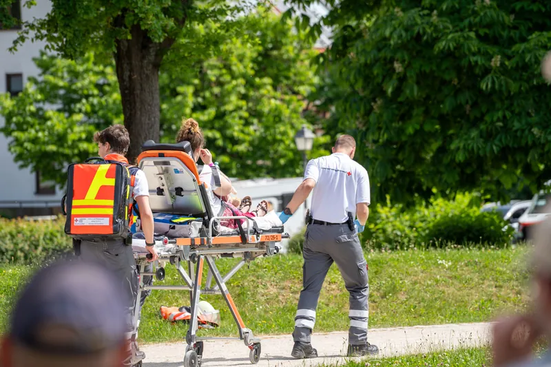 Helfer schieben eine Trage über den Weg, Zuschauer am Rand