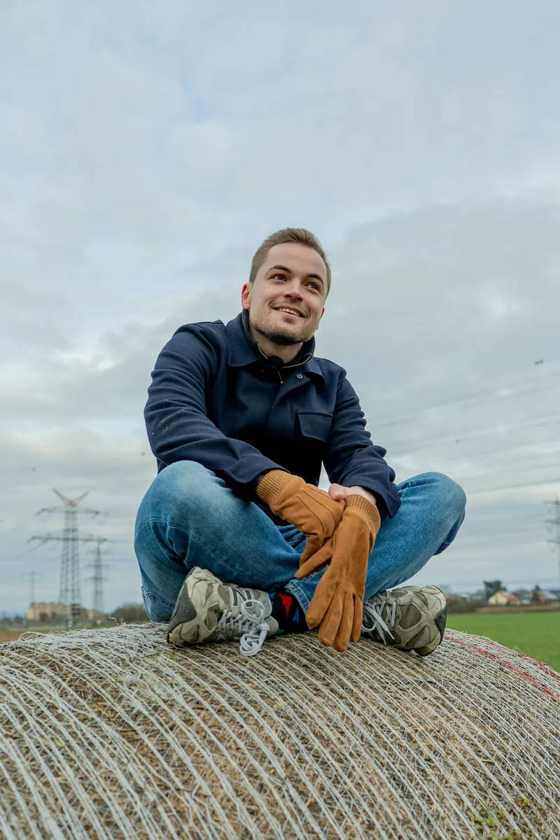 Junger Mann mit Arbeitshandschuhen sitzt auf einem Heuballen am Feldrand
